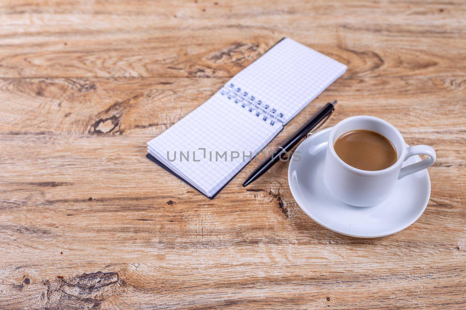 Small coffee cup on a saucer stands on a wooden table next to a notebook and pen by galinasharapova
