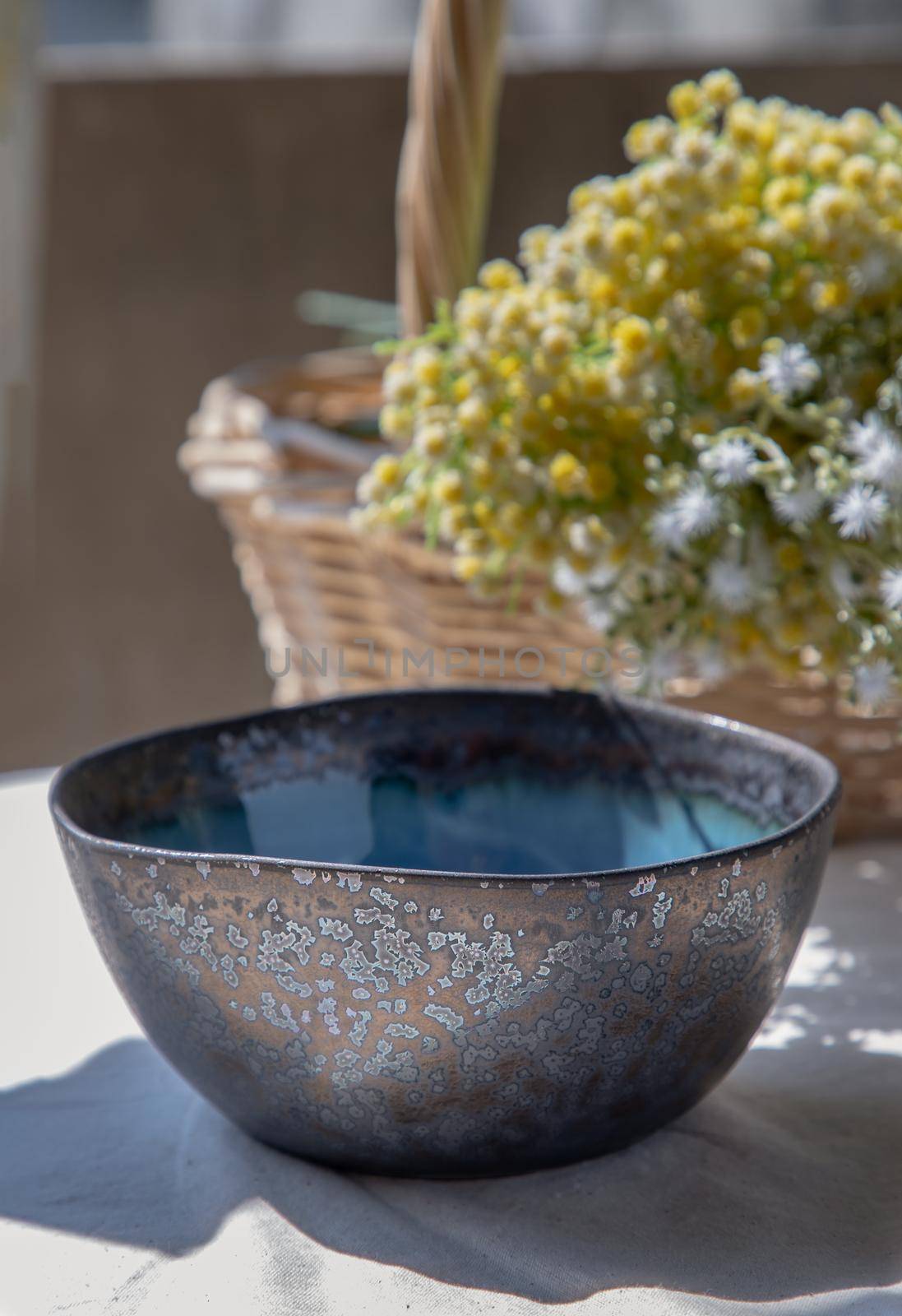 Ceramic bowl and bouquet of flowers in woven basket on white textured table cloth. Selective focus.