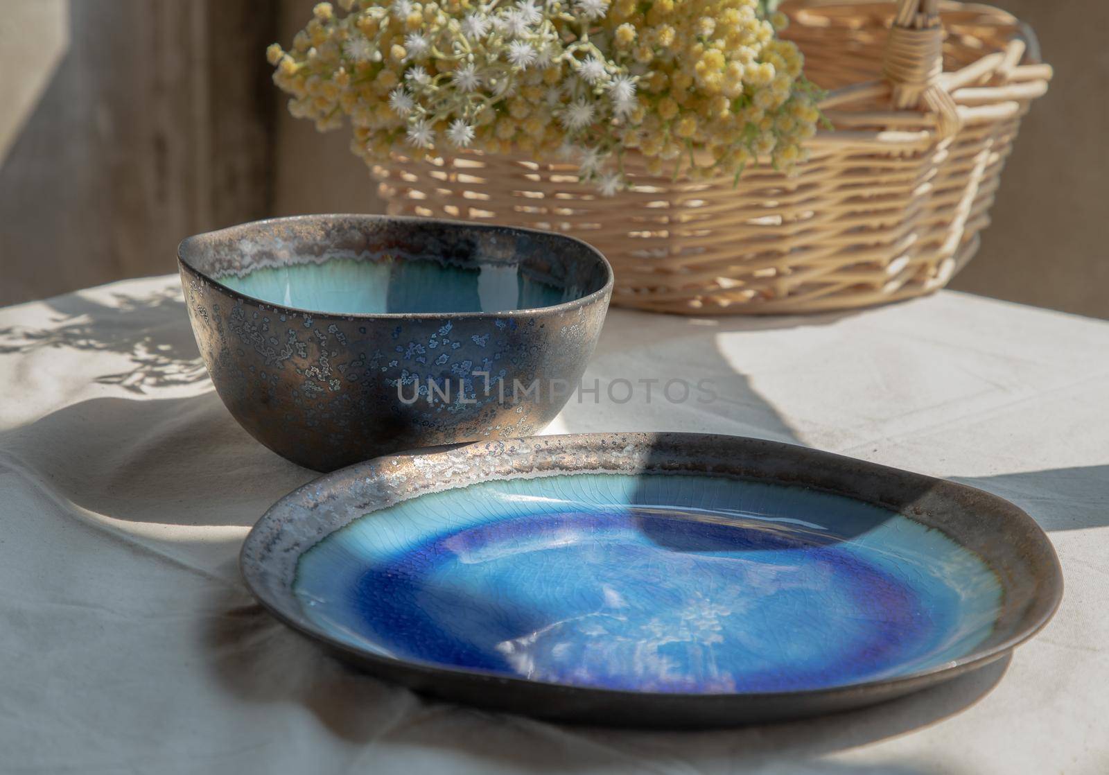 Set of Ceramic dishware and bouquet of flowers in woven basket on white textured table cloth. Ceramic tableware, Selective focus.