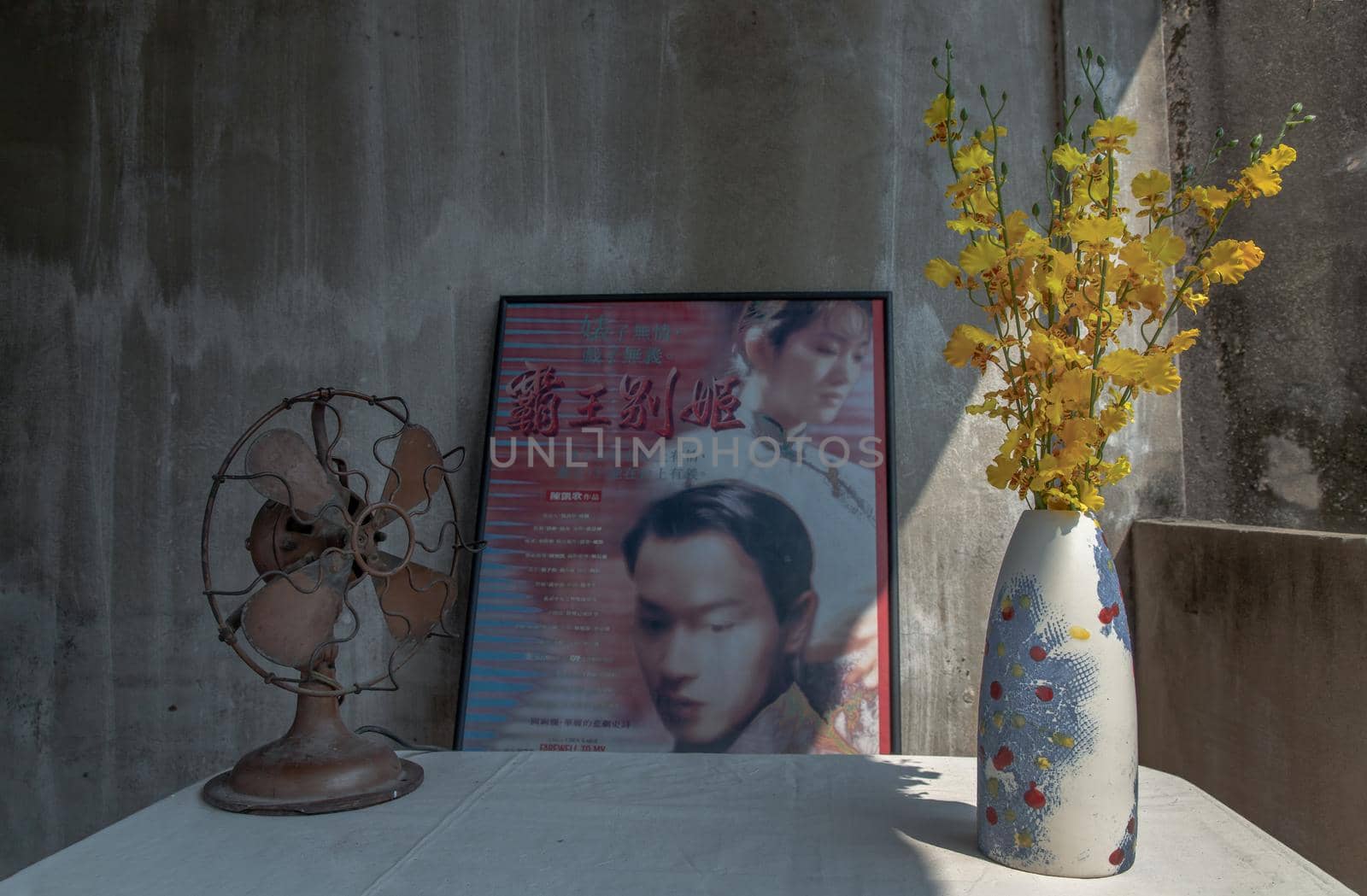 Yellow flowers in Handmade ceramic vase and Old vintage brown metal fan on white textured table cloth with Classic chinese poster movie frame with old ruins cement wall. Home decor, Selective focus.