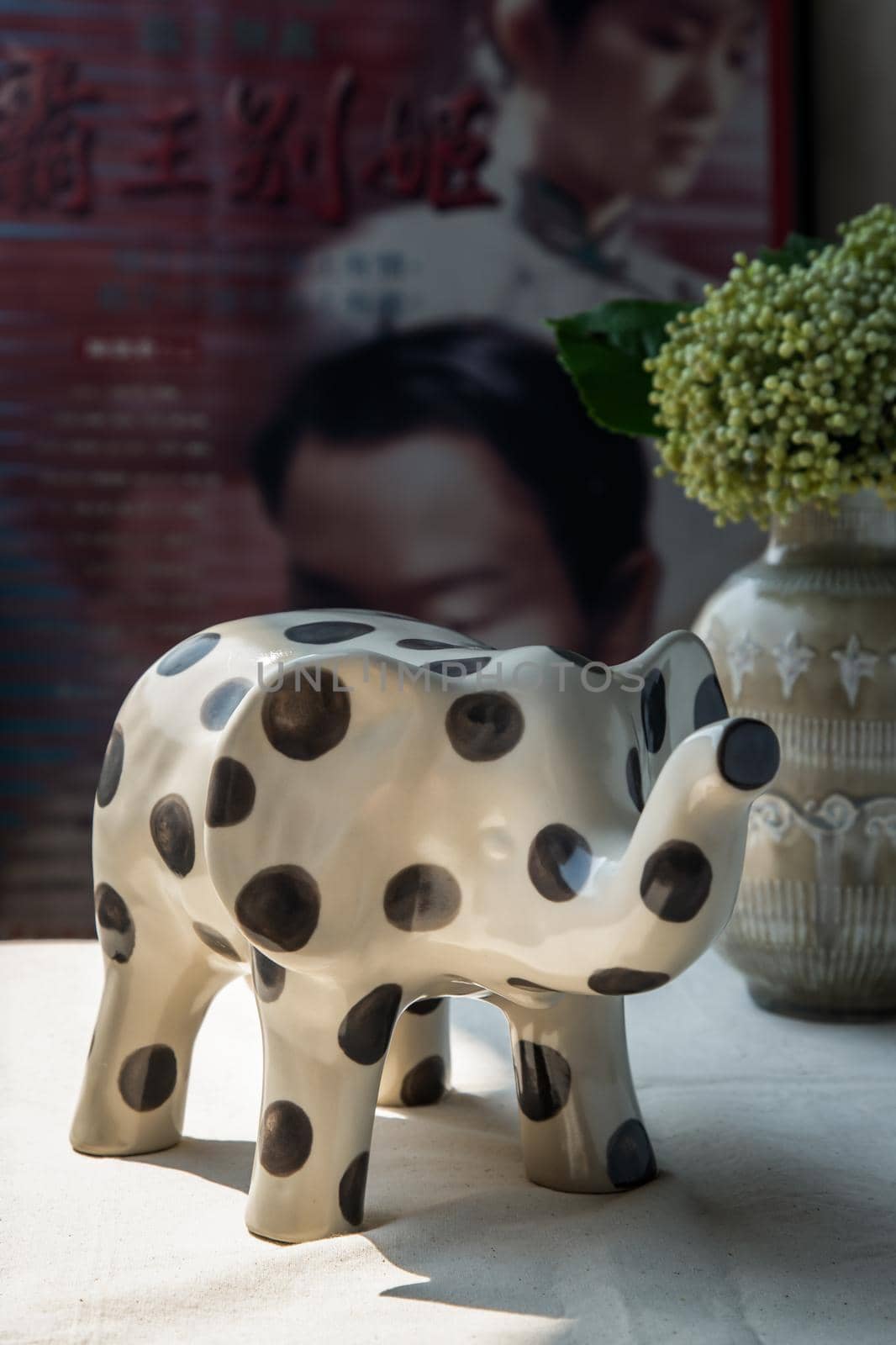 Bangkok, Thailand -Feb 06, 2021 :  Elephants shaped ceramic and Bouquet of flowers in ceramic vase on white textured table cloth in front of Classic chinese poster movie frame. Home decor, No focus, specifically.