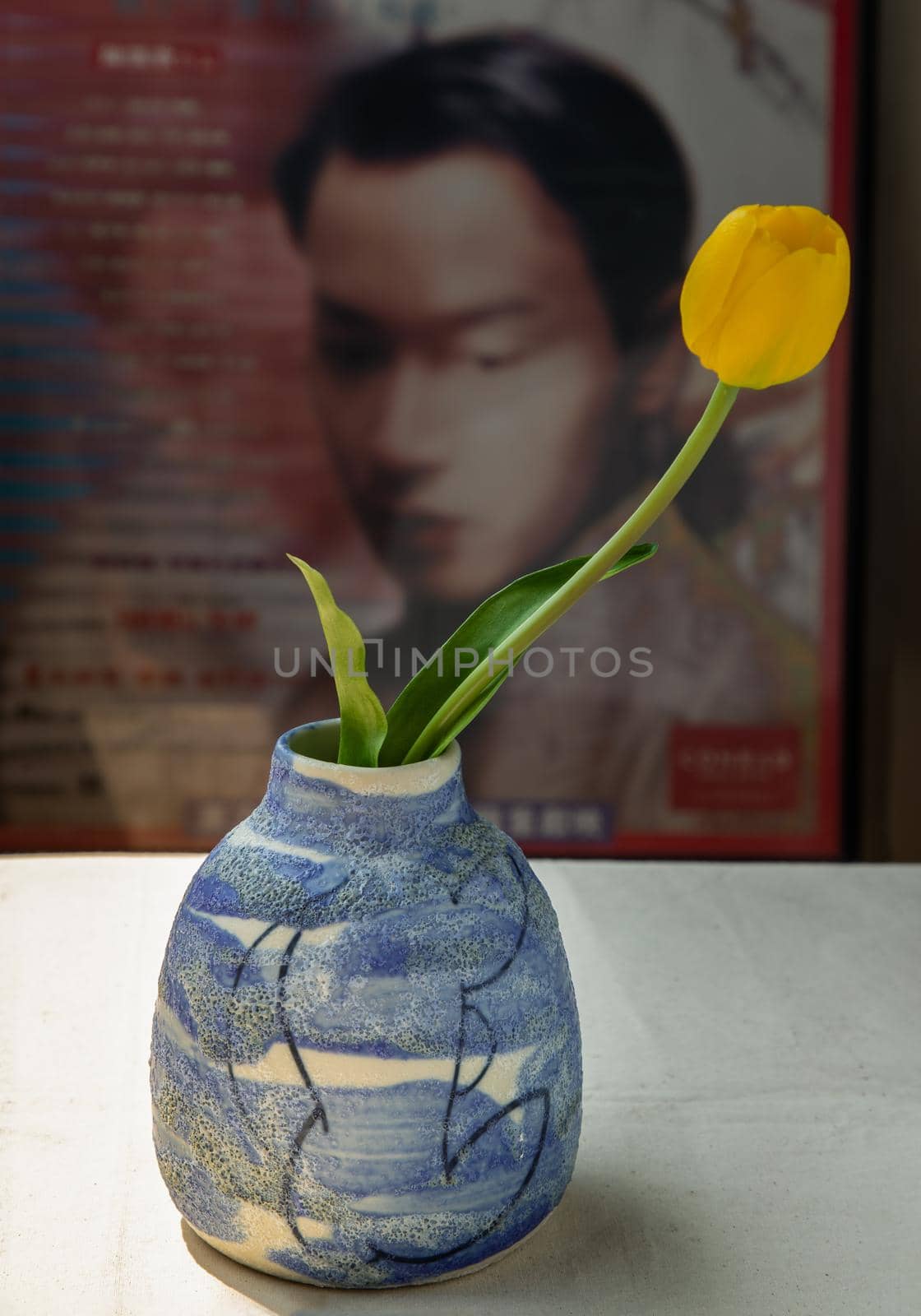 Bangkok, Thailand -Feb 06, 2021 : Yellow flower in Blue ceramic vase on white textured table cloth in front of Classic chinese poster movie frame. Home decor, Selective focus.
