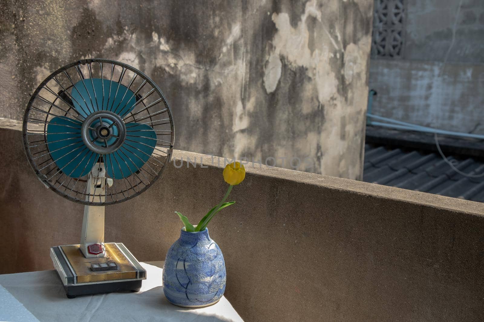 Yellow flower in Blue ceramic vase and old vintage brown metal fan on white textured table cloth with old cement wall at the balcony house. Home decor, Selective focus.