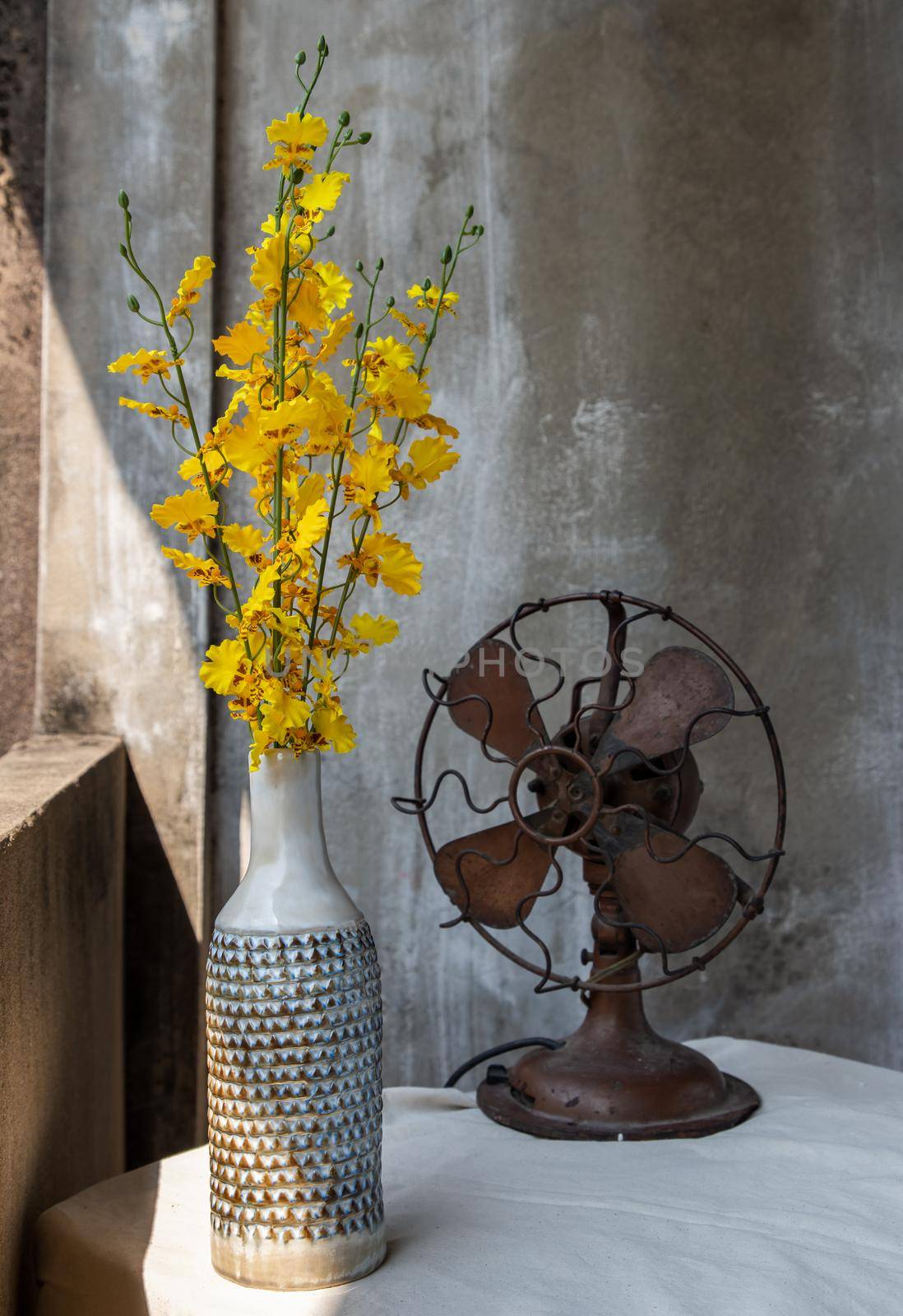 Yellow bouquet of flowers in White ceramic vase bottle shape and Vintage brown metal fan on white textured table cloth with old cement wall at the balcony house. Home decor, Selective focus.