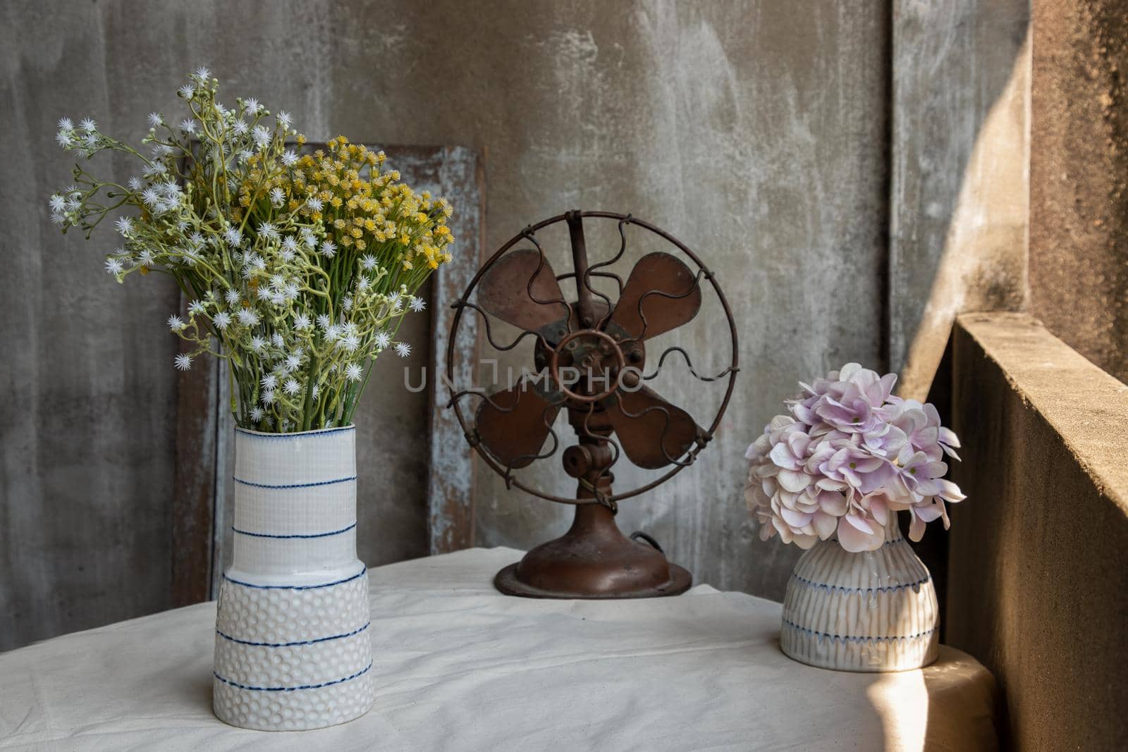 Bouquet of flowers in Two white handmade ceramic vase and Vintage brown metal fan on white textured table cloth with old cement wall at the balcony house. Home decor, Selective focus.