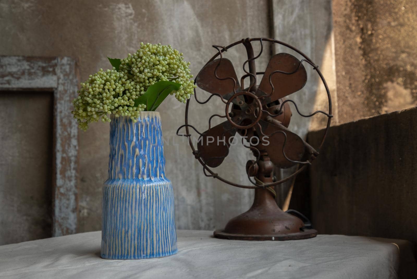 Bouquet of green flowers in Blue ceramic vase and Vintage brown metal fan on white textured table cloth with old cement wall at the balcony house. Home decor, Selective focus.