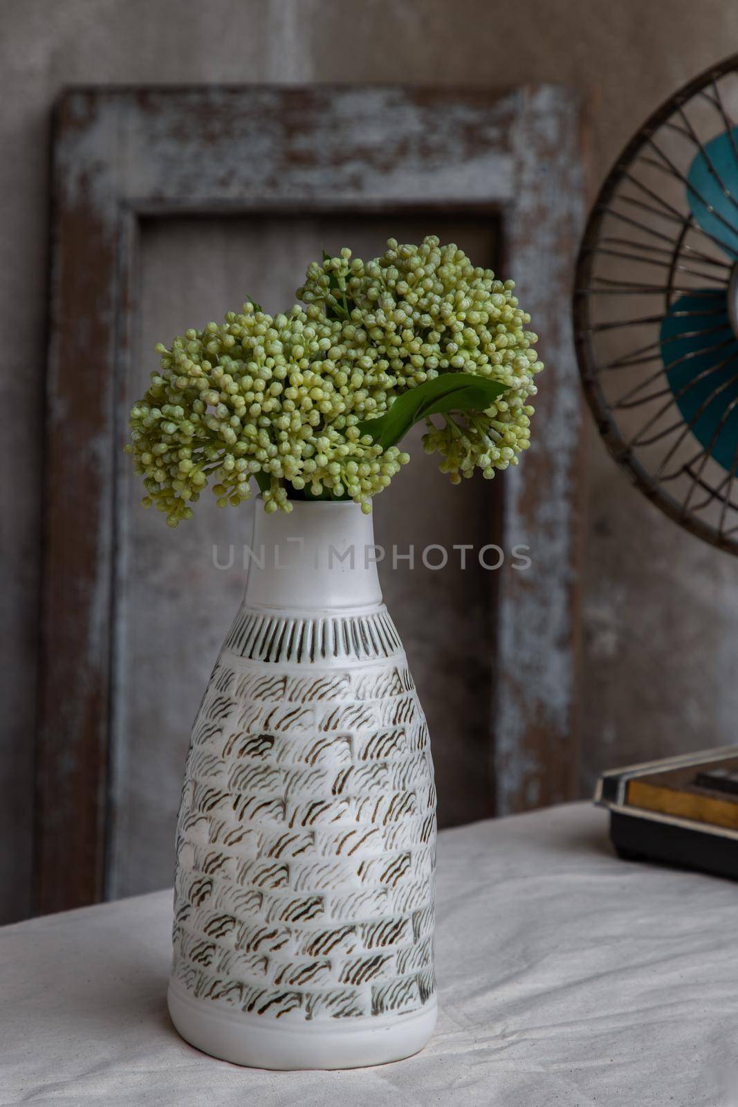 Bouquet of green flowers in White handmade ceramic vase bottle shape and Vintage fan on white textured table cloth with old cement wall. Home decor, Selective focus.