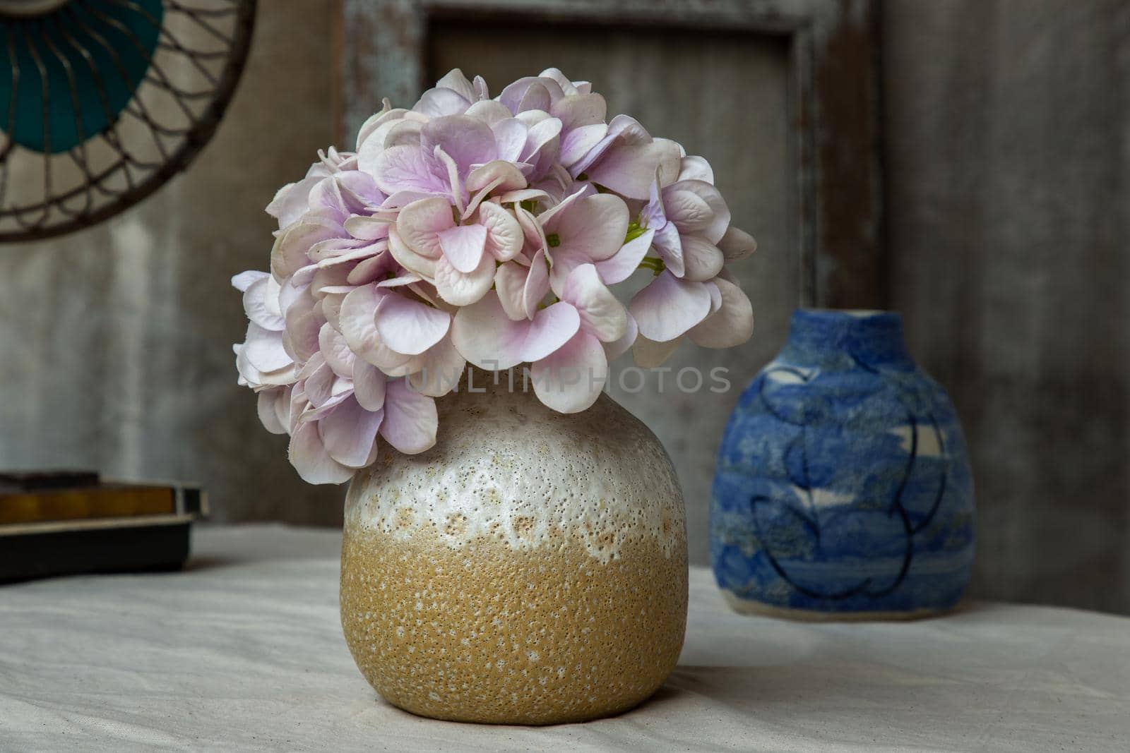 Bouquet of purple flowers in White with yellow handmade ceramic vase and Vintage fan on white textured table cloth with old cement wall. Home decor, Selective focus.