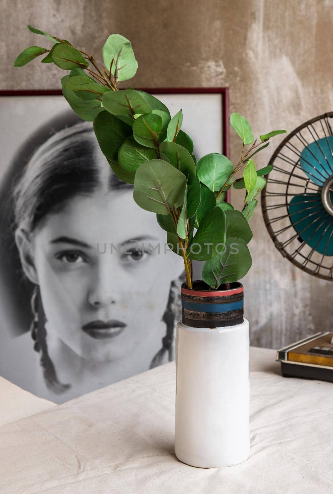 Bangkok, Thailand - Feb 06, 2021 : Green leaf in White ceramic vase and old vintage fan on white textured table cloth in front of Classic chinese poster movie frame with old cement wall. Home decor, Selective focus.