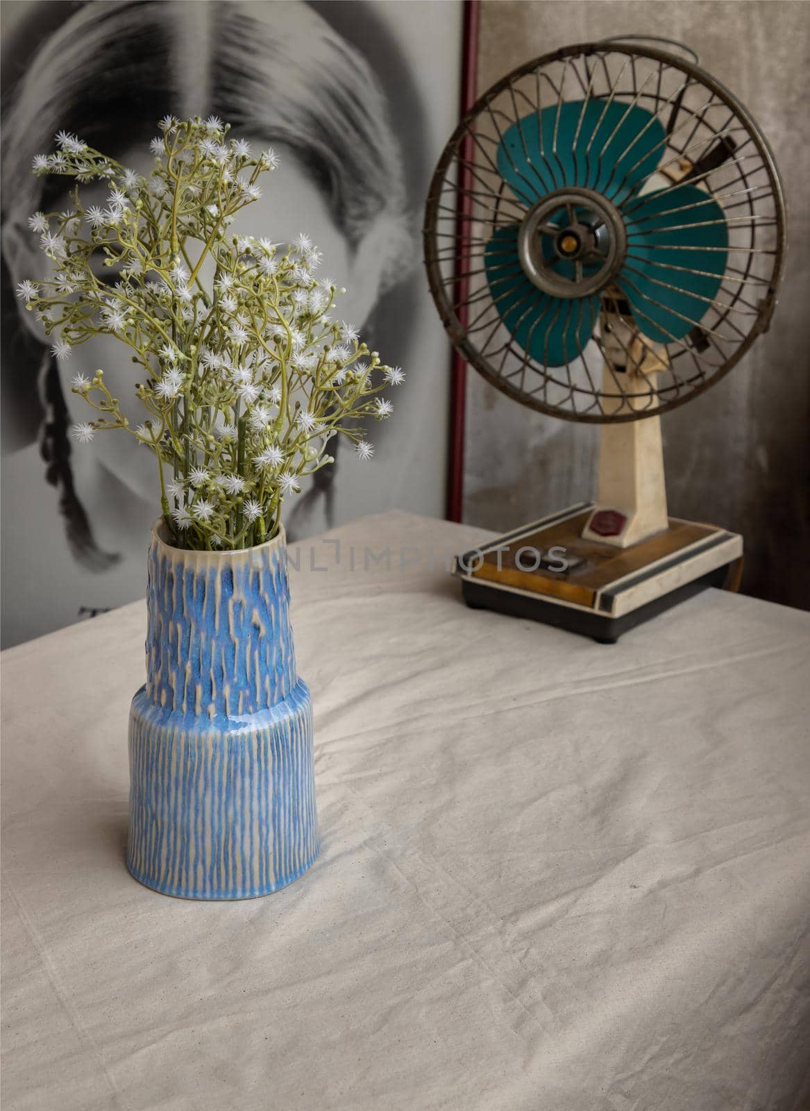 Bouquet of white flowers in Blue handmade ceramic vase and old vintage fan on white textured table cloth in front of Classic chinese poster movie frame with old cement wall. Home decor, Selective focus.