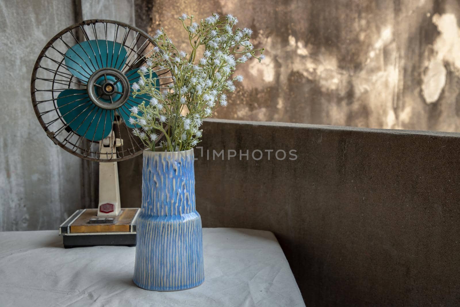 Bouquet of white flowers in Blue handmade ceramic vase and old vintage fan on white textured table cloth with old cement wall at the balcony house. Home decor, Copy space, Selective focus.