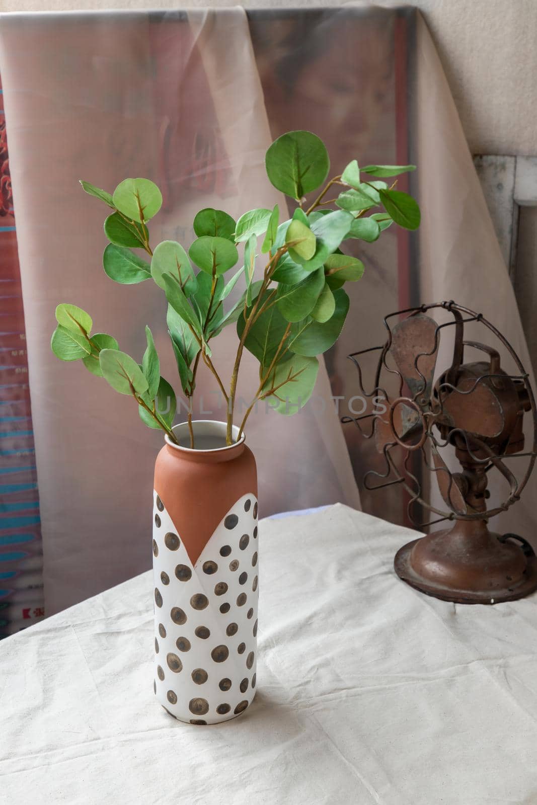 Green leaf in Handmade ceramic vase and Vintage fan on white textured table cloth with old cement wall. Home decor, Selective focus.