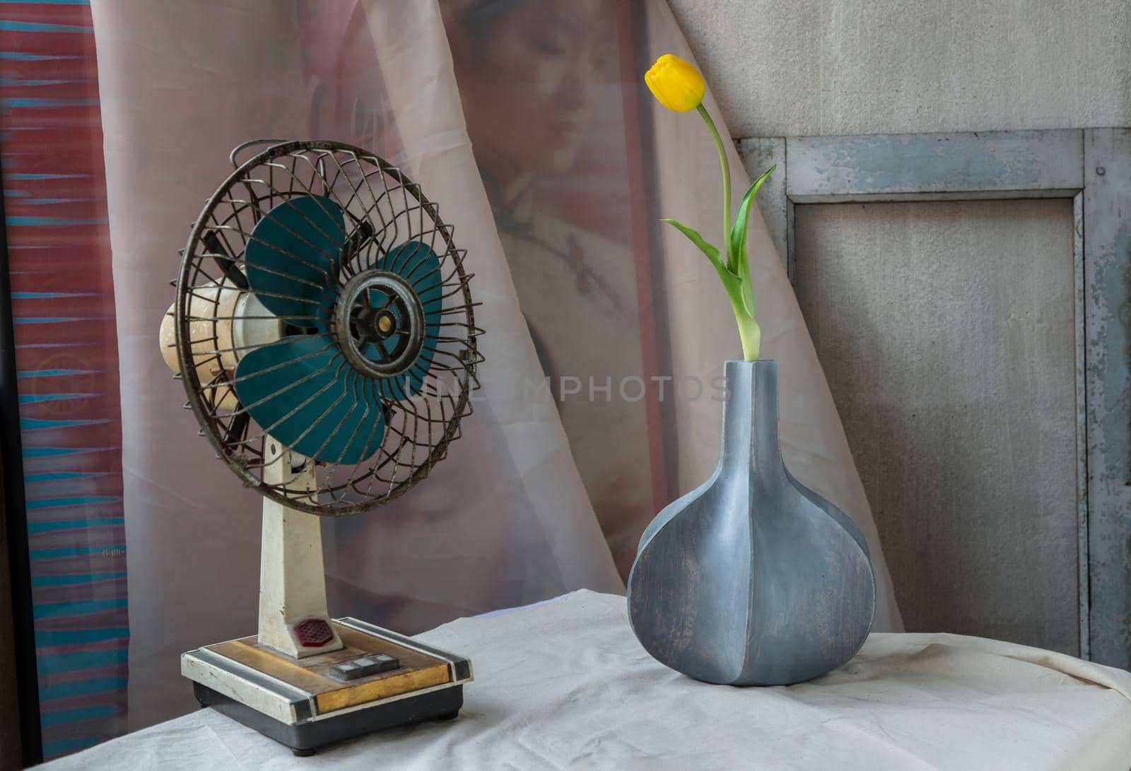Yellow flower in Blue wooden vase and Vintage fan on white textured table cloth with old cement wall. Home decor, Selective focus.