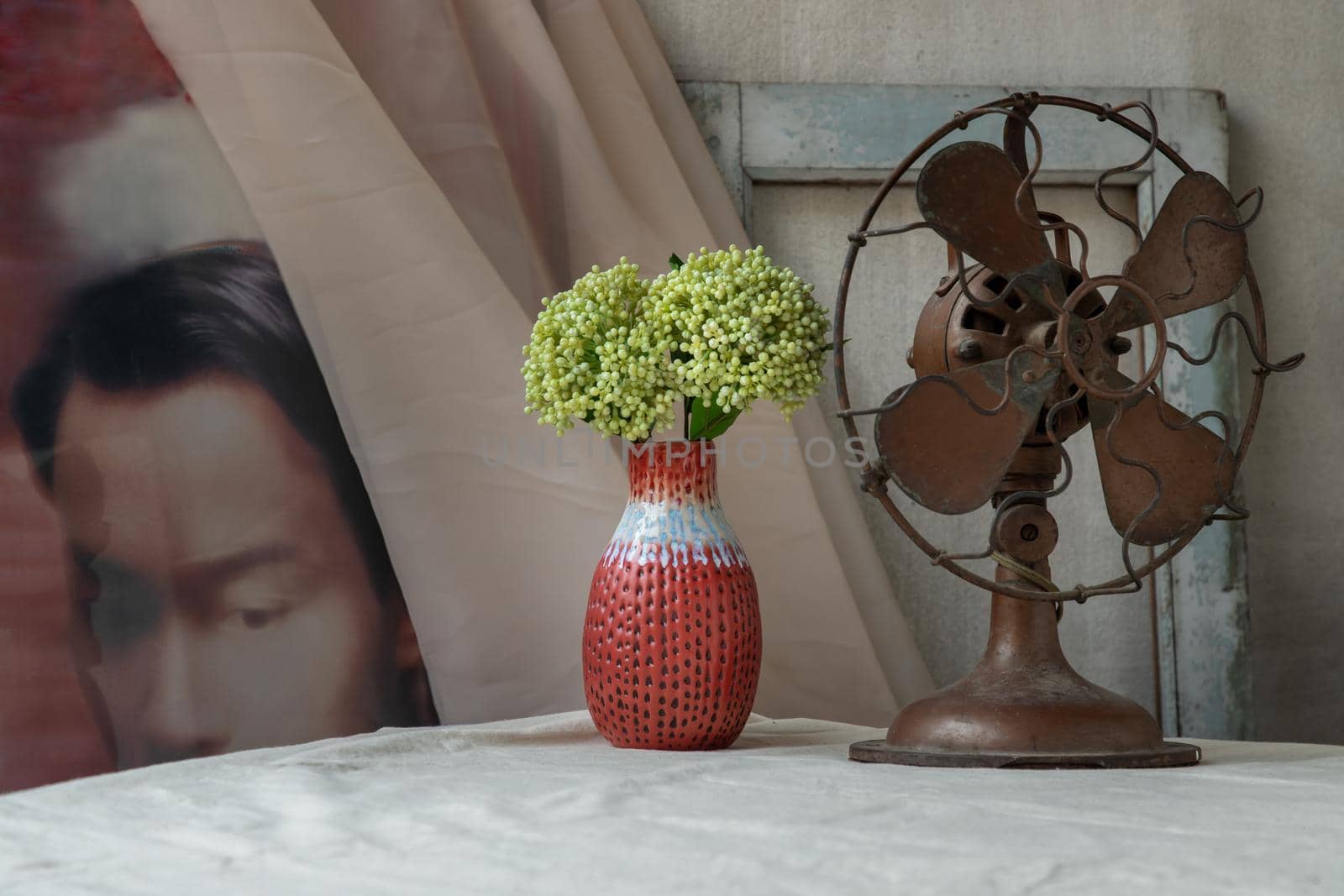 Bangkok, Thailand - Feb 06, 2021 : Bouquet of green flowers in Red handmade ceramic vase and old vintage fan on white textured table cloth in front of Classic chinese poster movie frame with old cement wall. Home decor, Selective focus.