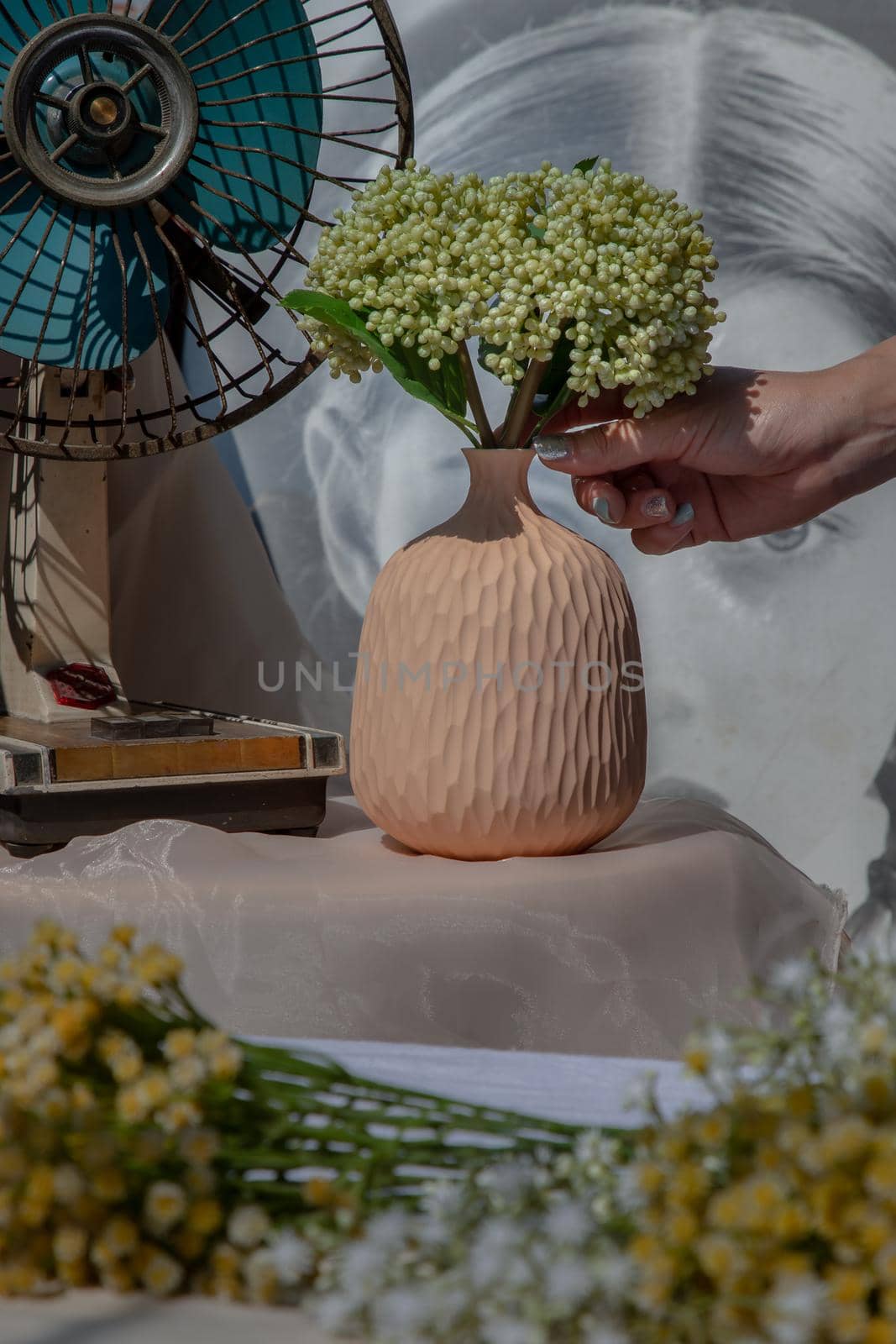 Hands of a young woman with Bouquet of green flowers in Handmade ceramic vase and old vintage fan on pink textured table cloth in front of Classic chinese poster movie frame. Home decor, Selective focus.