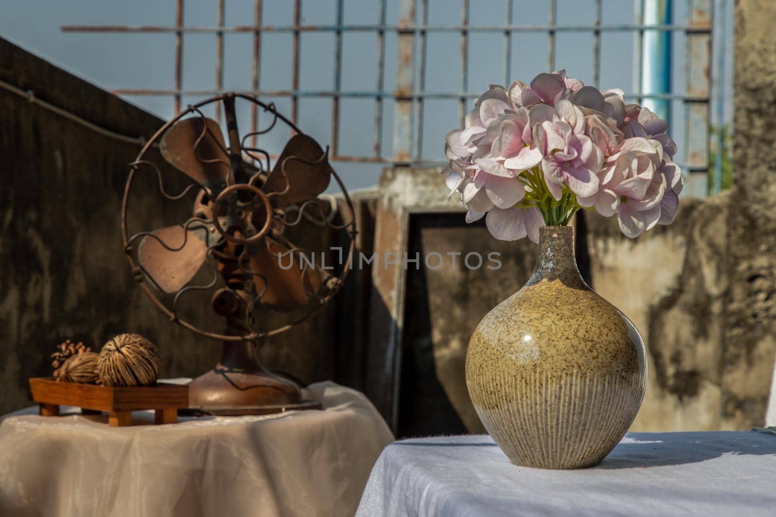 Bouquet of purple flowers in Handmade ceramic vase on white textured table cloth and vintage fan with Old cement wall at the balcony house. Home decor, Selective focus.