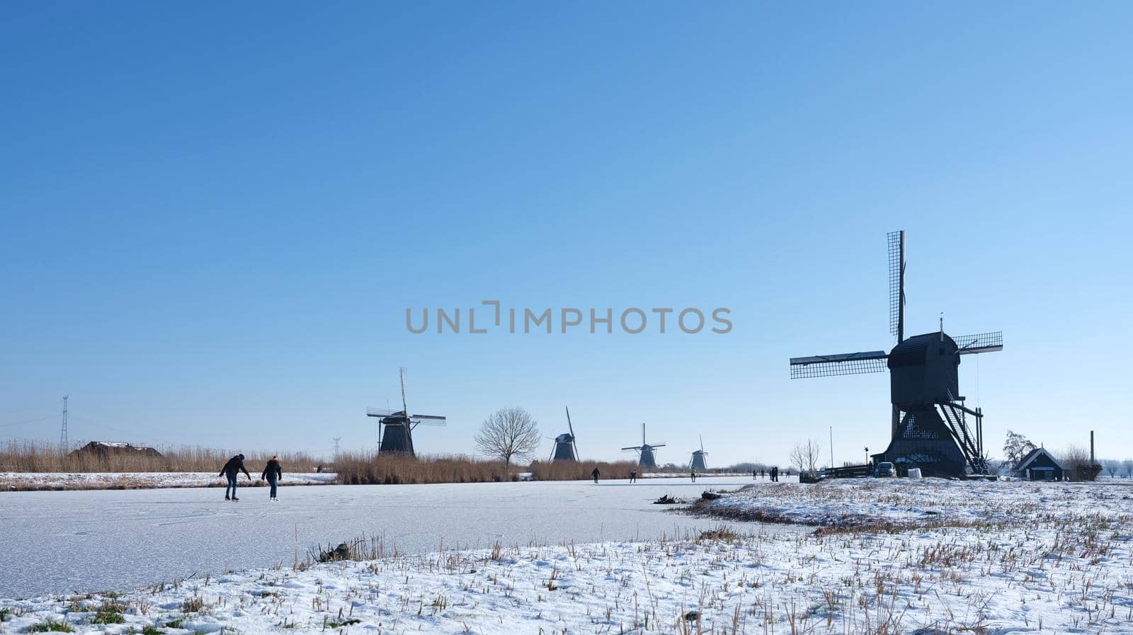 people skate on the ice of canal near kinderdijk with a lot of windmills in holland on sunny winter day