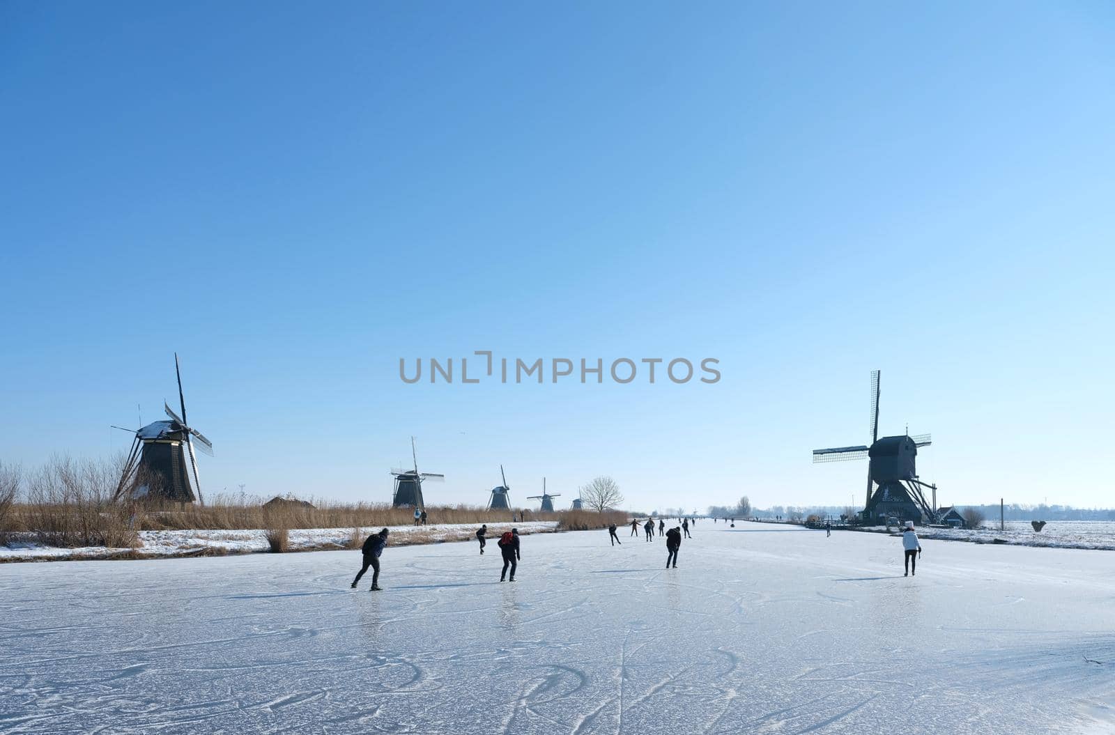 people skate on the ice of canal near kinderdijk with a lot of windmills in holland on sunny winter day