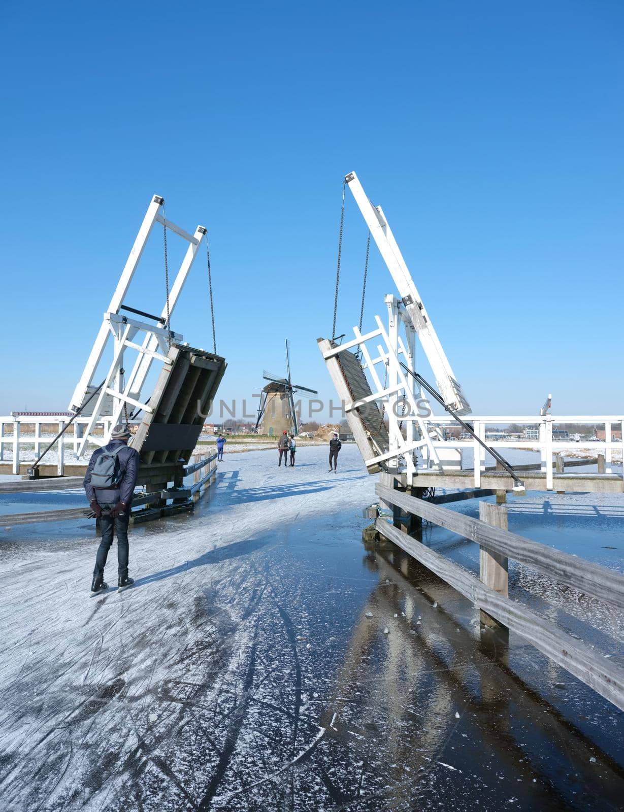 people skate through drawbridge in kinderdijk on frozen water of canal on sunny winter day in holland by ahavelaar