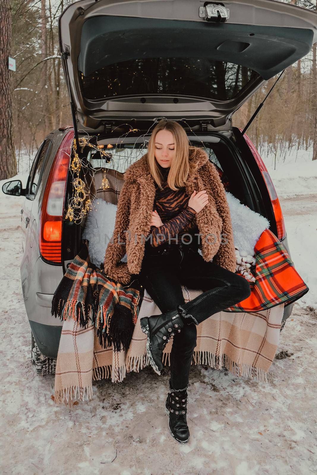 Blonde woman wrapped in blanket in trunk car. Travel in winter. Car decorated with festive Christmas lights. Outdoor picnic. Unity with nature