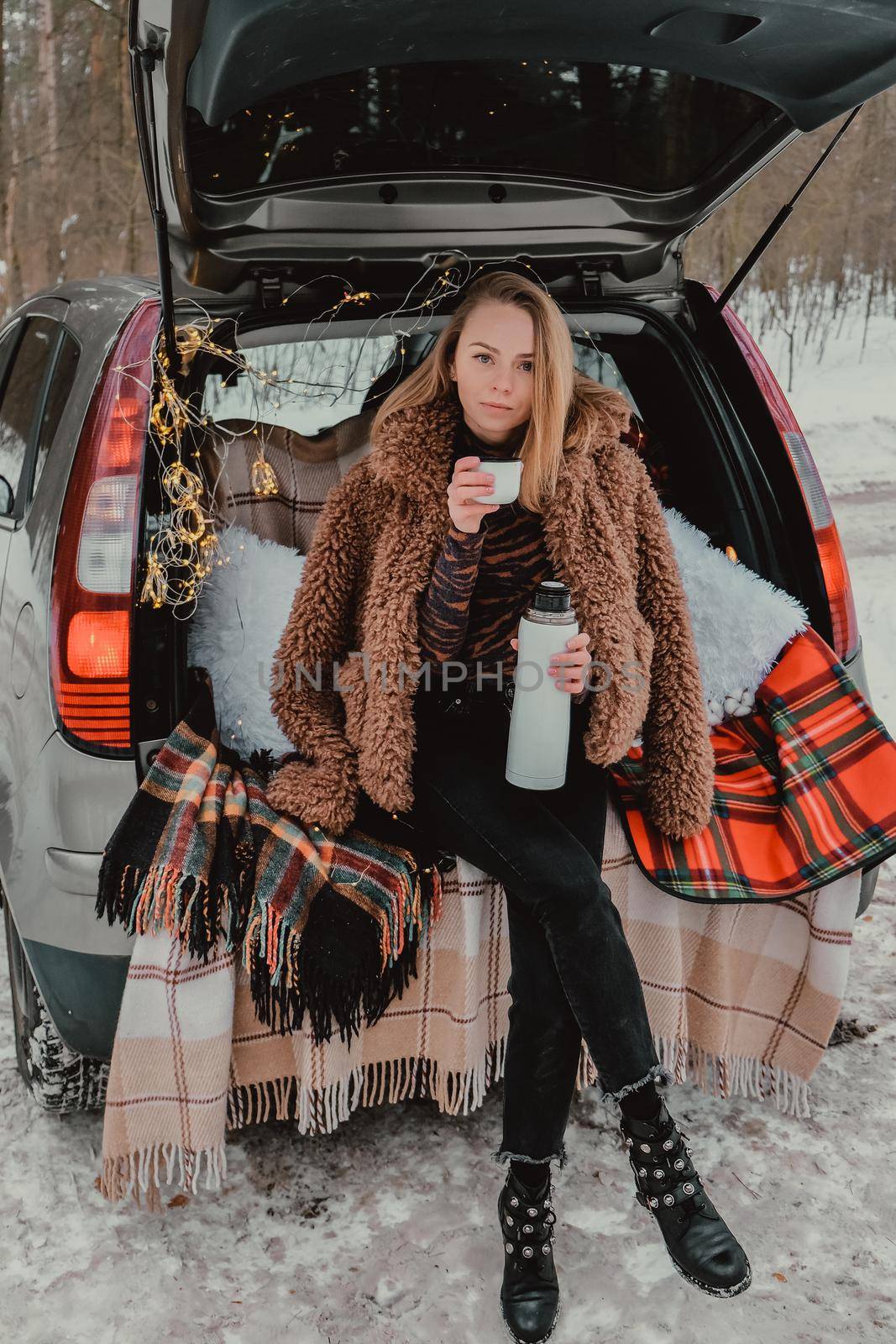 Blonde woman wrapped in blanket in trunk car drinking coffee tea from thermos. Travel in winter. Car decorated with festive Christmas lights. Outdoor picnic. Unity with nature