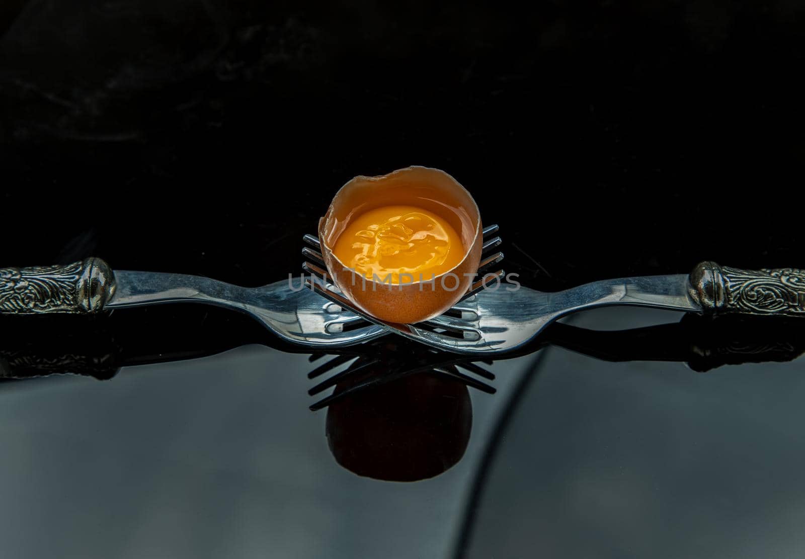 Broken fresh chicken egg (Hen egg) balanced on a composition of two intertwined forks over black table. Copy space, Selective focus.