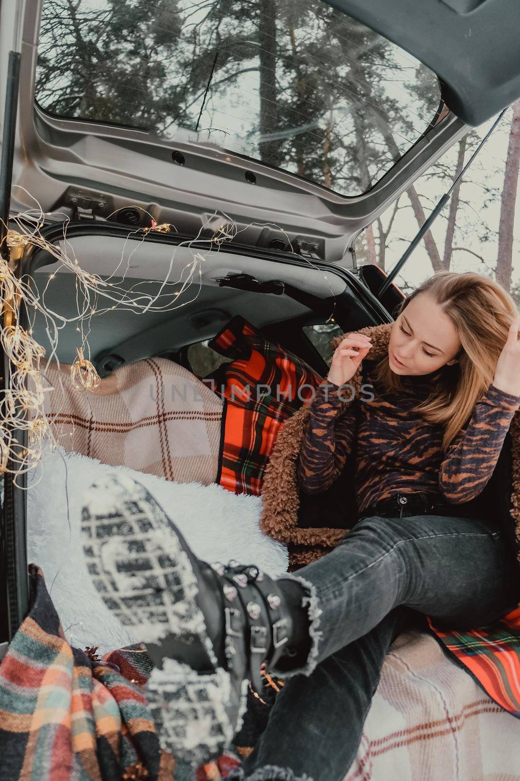 Blonde woman wrapped in blanket in trunk car. Travel in winter. Car decorated with festive Christmas lights. Outdoor picnic. Unity with nature