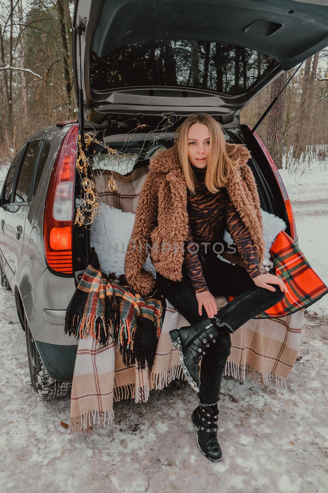 Blonde woman wrapped in blanket in trunk car. Travel in winter. Car decorated with festive Christmas lights. Outdoor picnic. Unity with nature