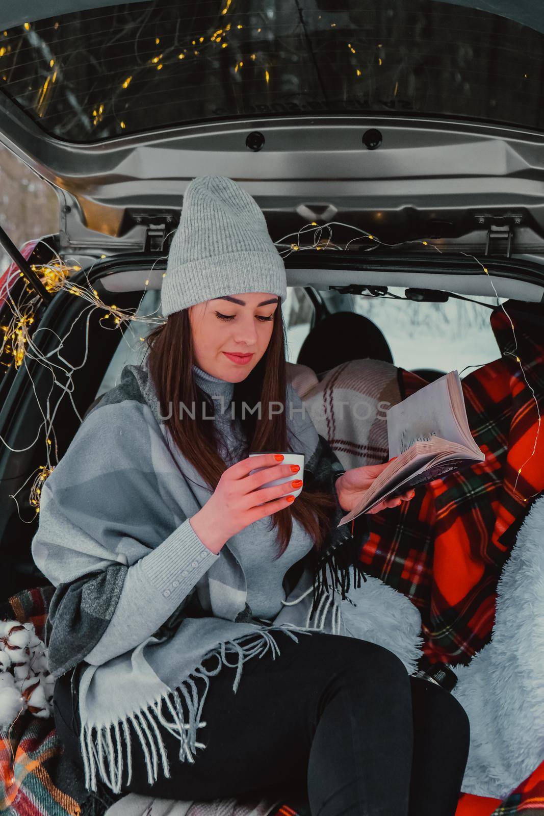 Brunette woman wrapped in blanket in trunk car drinking coffee tea from thermos. Travel in winter. Car decorated with festive Christmas lights. Outdoor picnic. Unity with nature