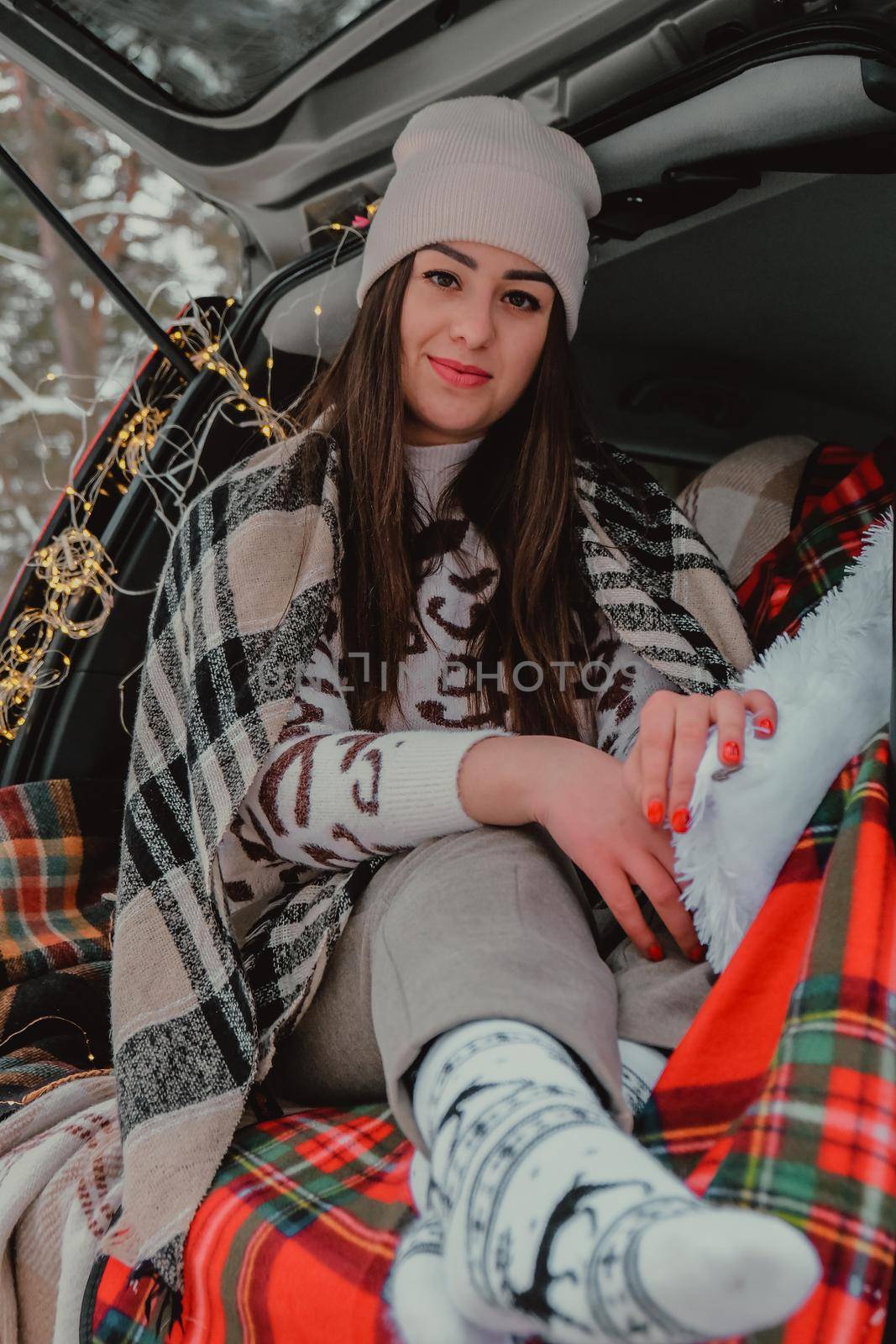 Brunette woman wrapped in blanket in trunk car drinking coffee tea from thermos. Travel in winter. Car decorated with festive Christmas lights. Outdoor picnic. Unity with nature