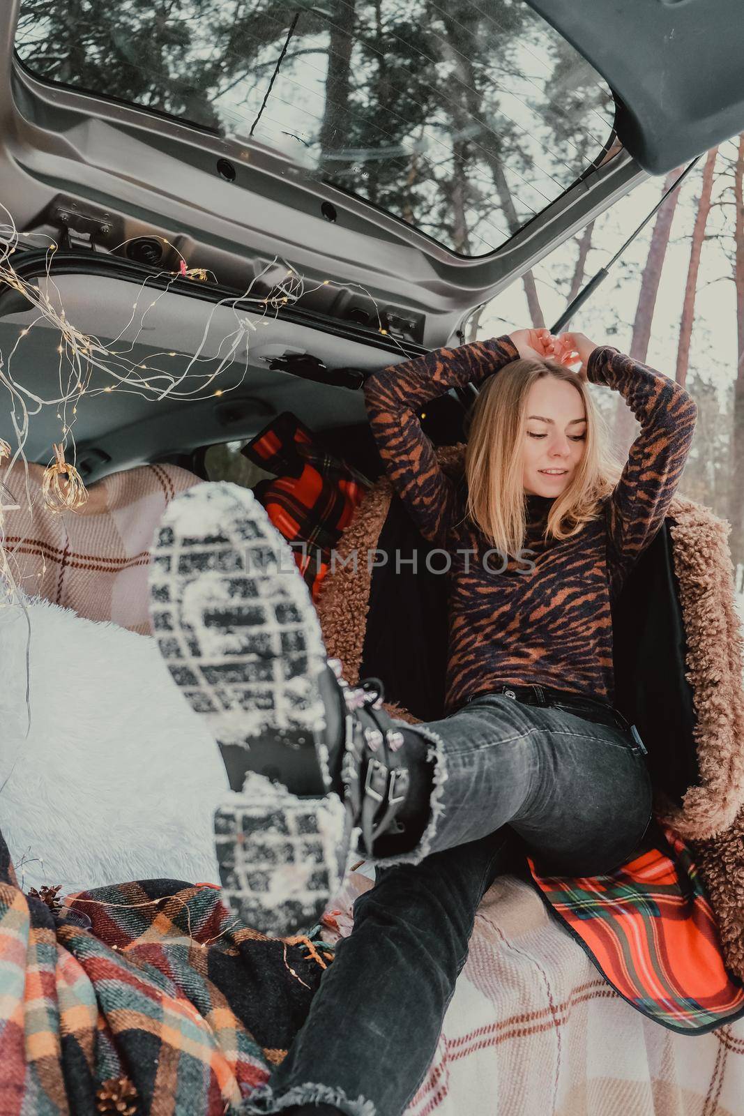 Blonde woman wrapped in blanket in trunk car. Travel in winter. Car decorated with festive Christmas lights. Outdoor picnic. Unity with nature