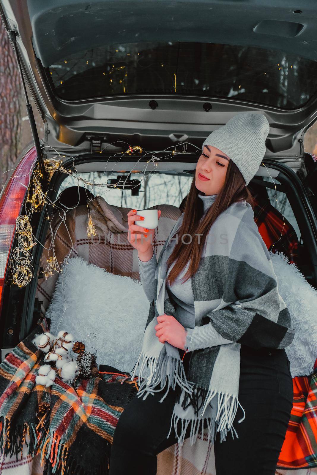 Brunette woman wrapped in blanket in trunk car drinking coffee tea from thermos. Travel in winter. Car decorated with festive Christmas lights. Outdoor picnic. Unity with nature