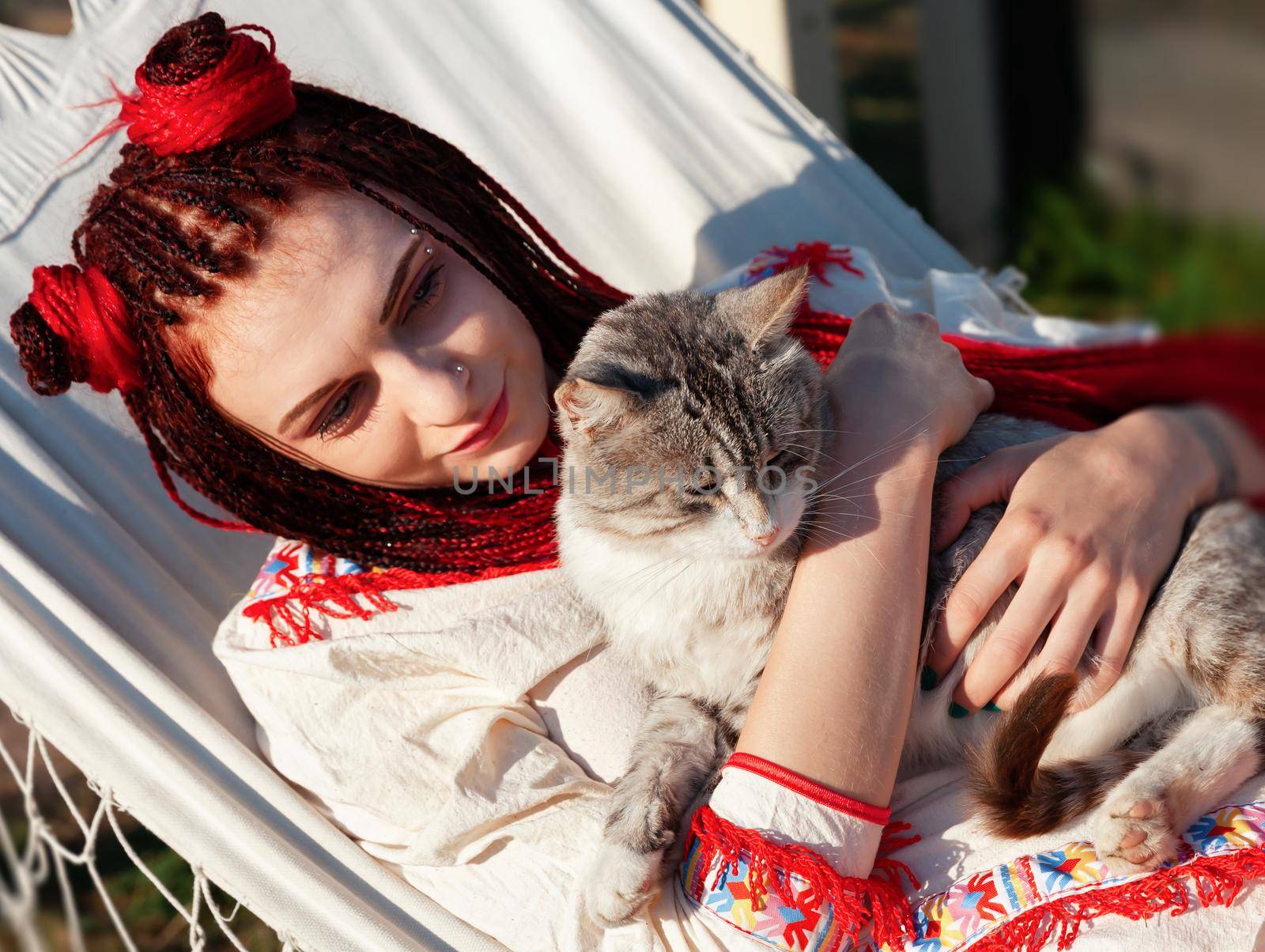 Young woman with scarlet dreadlocks in national dress lying in a hammock and playing with a cat. Outdoors portrait