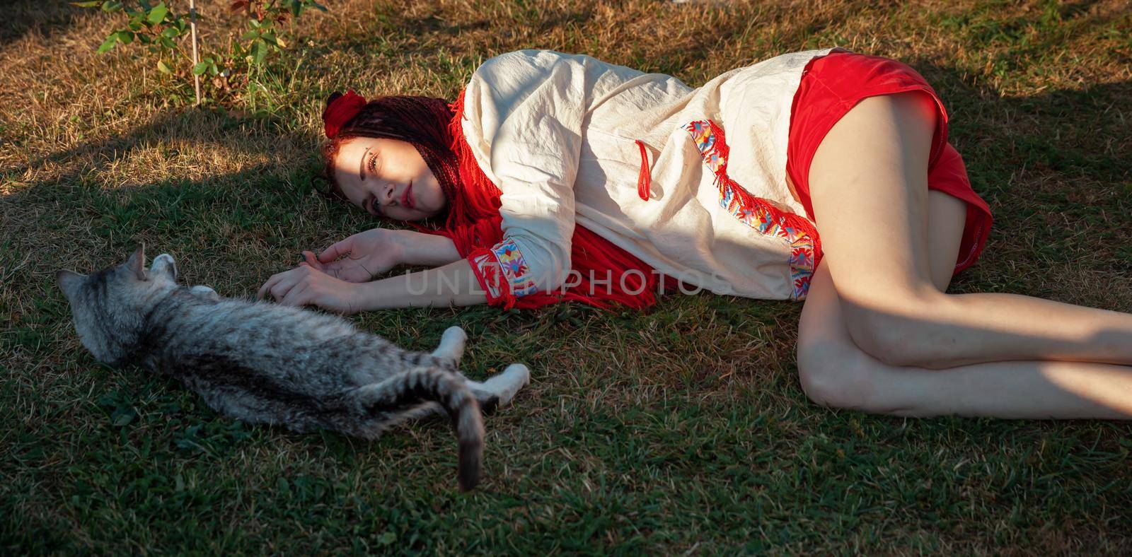 Young woman with scarlet dreadlocks in national dress lying on the grass and playing with the cat. Outdoors portrait