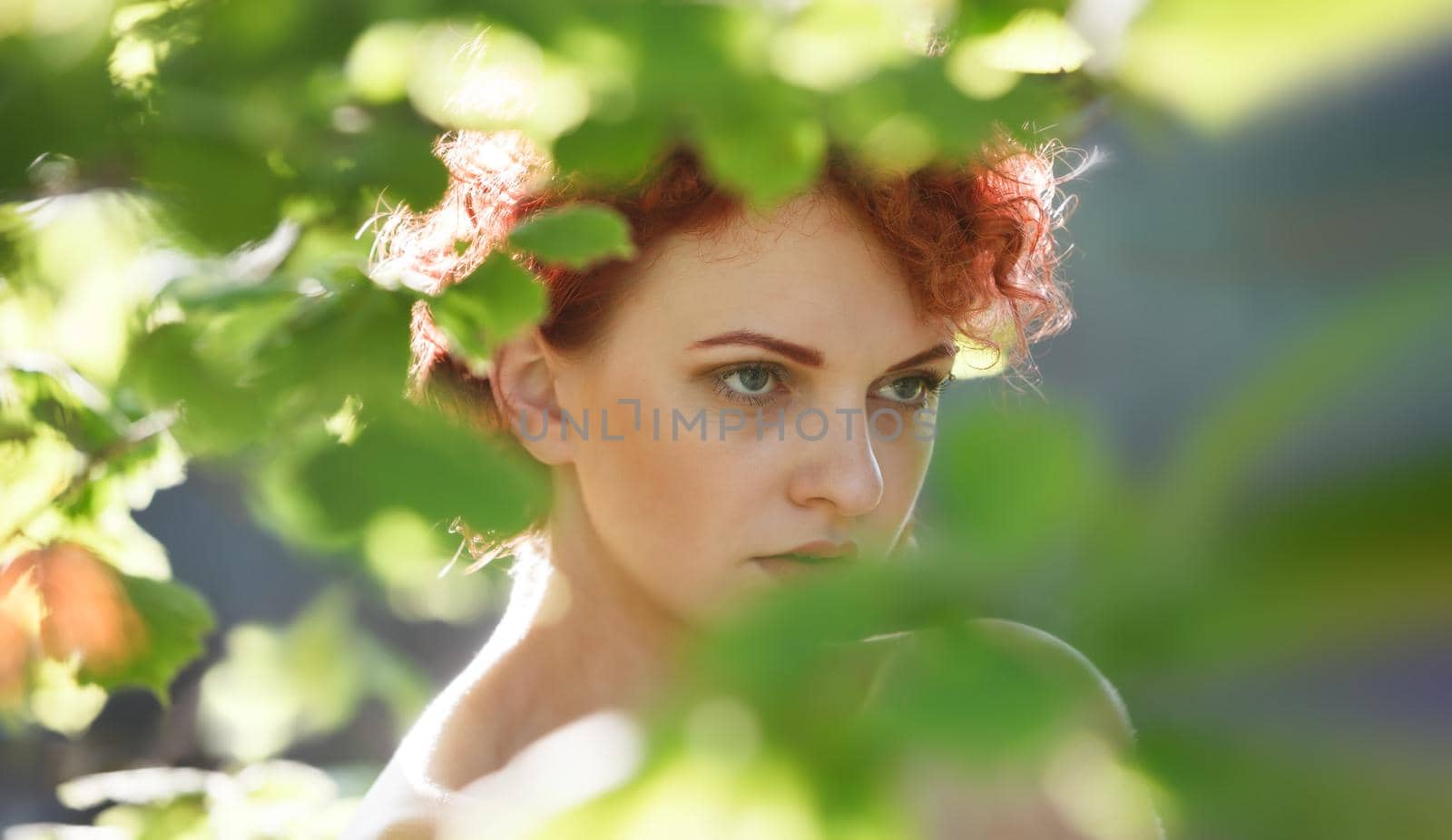 Portrait of a young red-haired woman among the foliage of trees illuminated by the suns rays