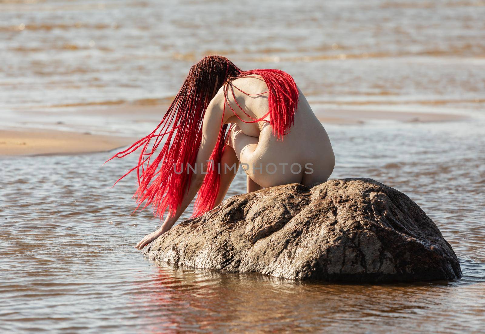 Beautiful girl outdoors enjoying nature. Young naked woman with scarlet dreadlocks on the stone in the sea enjoys the tranquility of nature