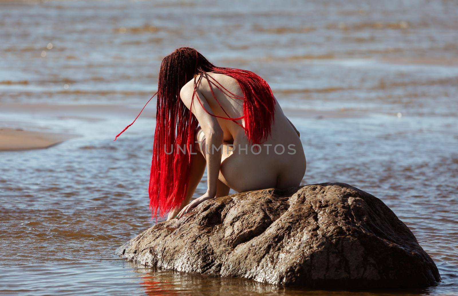 Beautiful girl outdoors enjoying nature. Young naked woman with scarlet dreadlocks on the stone in the sea enjoys the tranquility of nature
