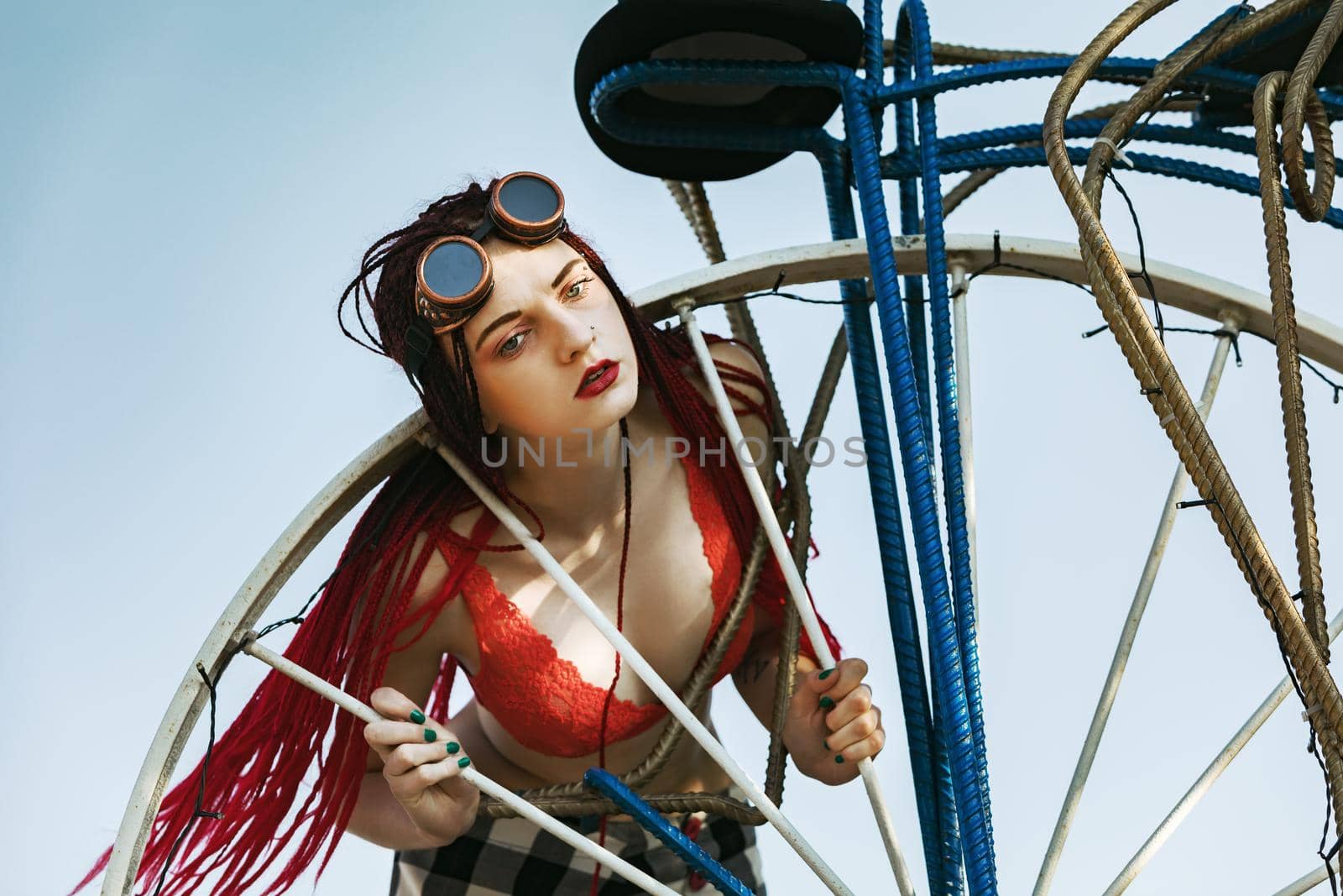 Glamorous girl with scarlet dreadlocks, a red swimsuit, black hat and welding glasses poses on the blue sky background. Free space for your text