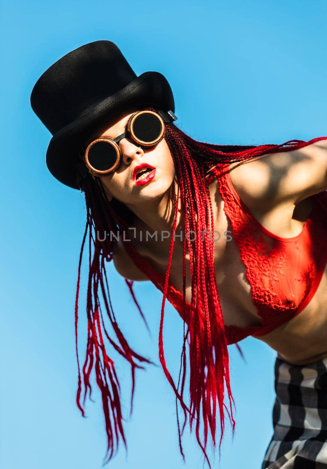 Glamorous girl with scarlet dreadlocks, a red swimsuit, black hat and welding glasses poses on the blue sky background. Free space for your text