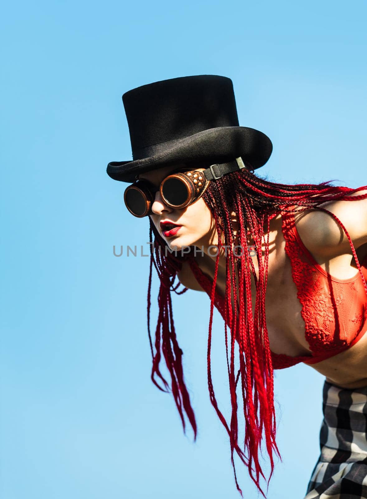 Glamorous girl with scarlet dreadlocks, a red swimsuit, black hat and welding glasses poses on the blue sky background. Free space for your text
