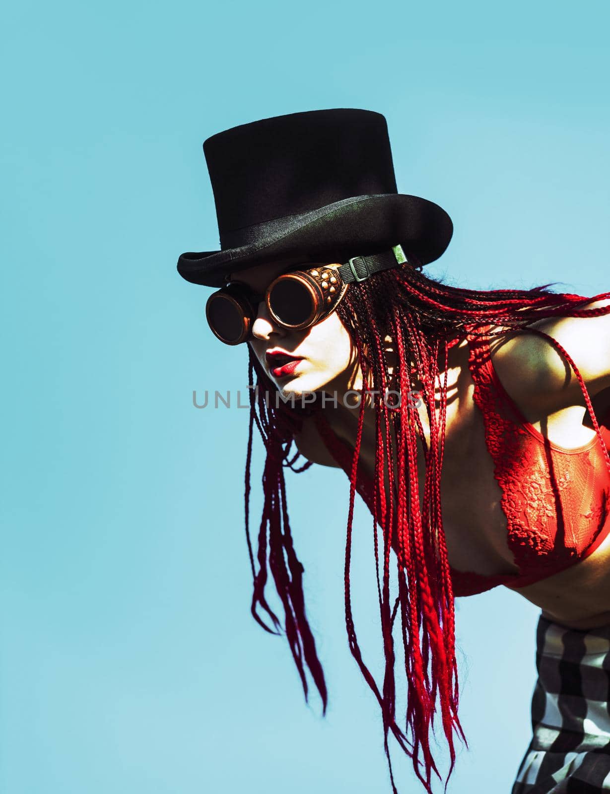 Glamorous girl with scarlet dreadlocks, a red swimsuit, black hat and welding glasses poses on the blue sky background. Free space for your text