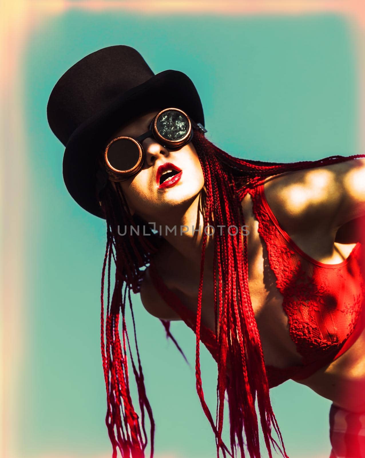 Glamorous girl with scarlet dreadlocks, a red swimsuit, black hat and welding glasses poses on the blue sky background. Free space for your text