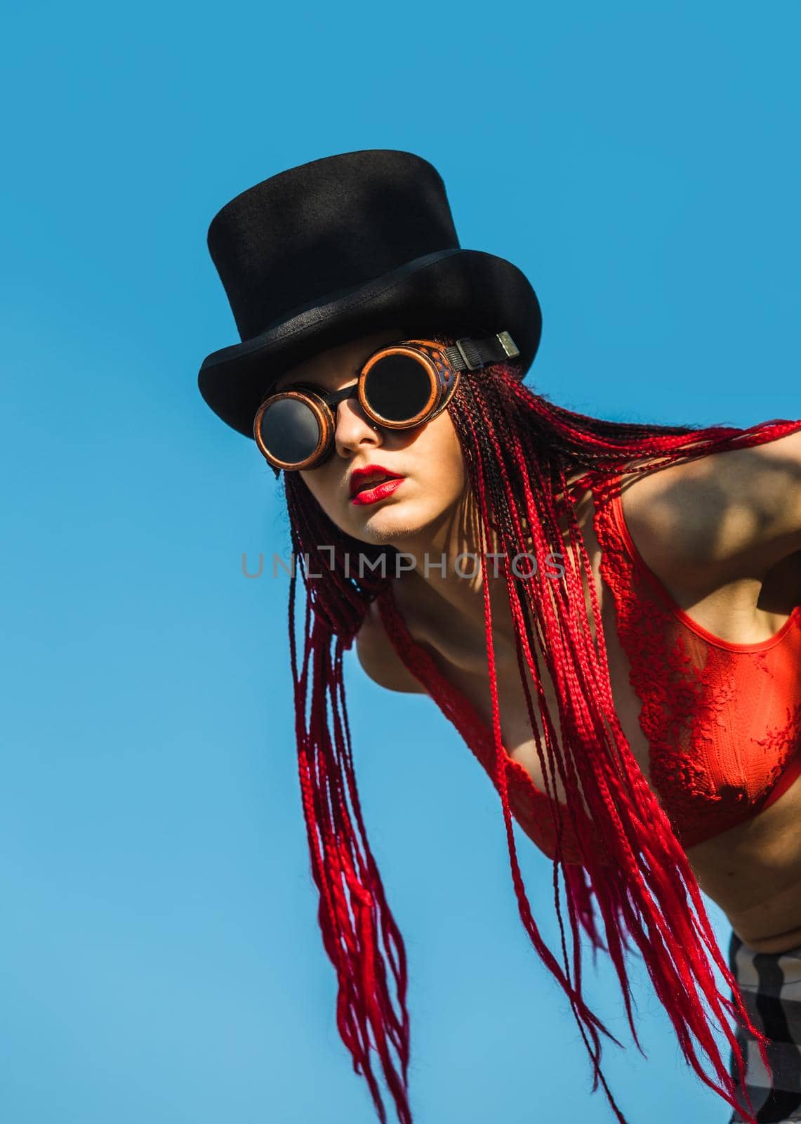 Glamorous girl with scarlet dreadlocks, a red swimsuit, black hat and welding glasses poses on the blue sky background. Free space for your text