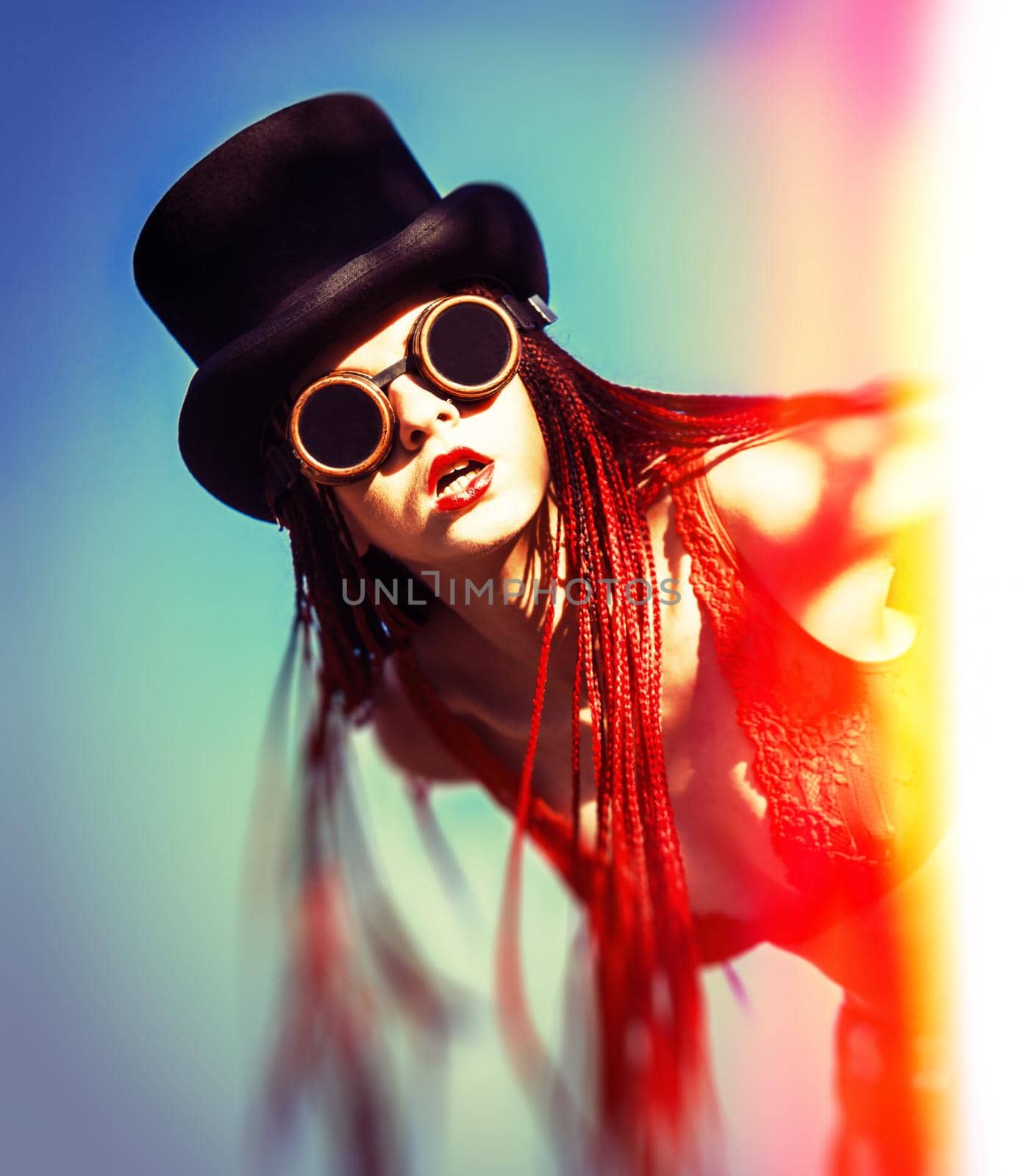 Glamorous girl with scarlet dreadlocks, a red swimsuit, black hat and welding glasses poses on the blue sky background. Free space for your text