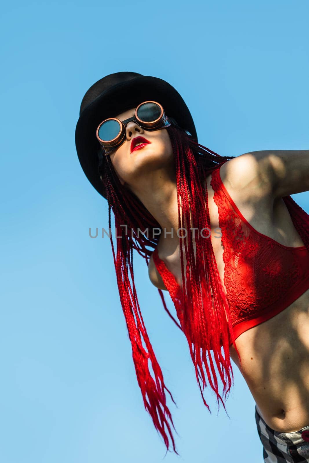 Glamorous girl with scarlet dreadlocks, a red swimsuit, black hat and welding glasses poses on the blue sky background. Free space for your text