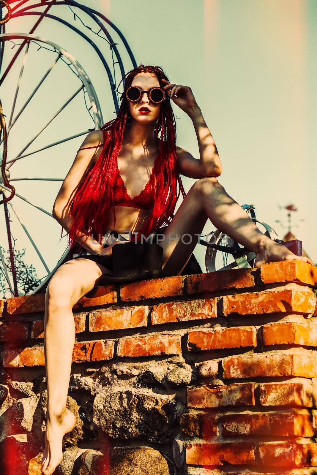 Glamorous girl with scarlet dreadlocks, a red swimsuit and welding glasses sits on a brick wall and poses on the blue sky background. Free space for your text