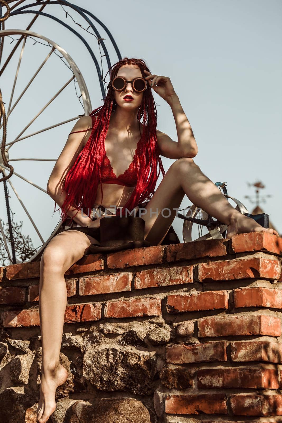 Glamorous girl with scarlet dreadlocks, a red swimsuit and welding glasses sits on a brick wall and poses on the blue sky background. Free space for your text