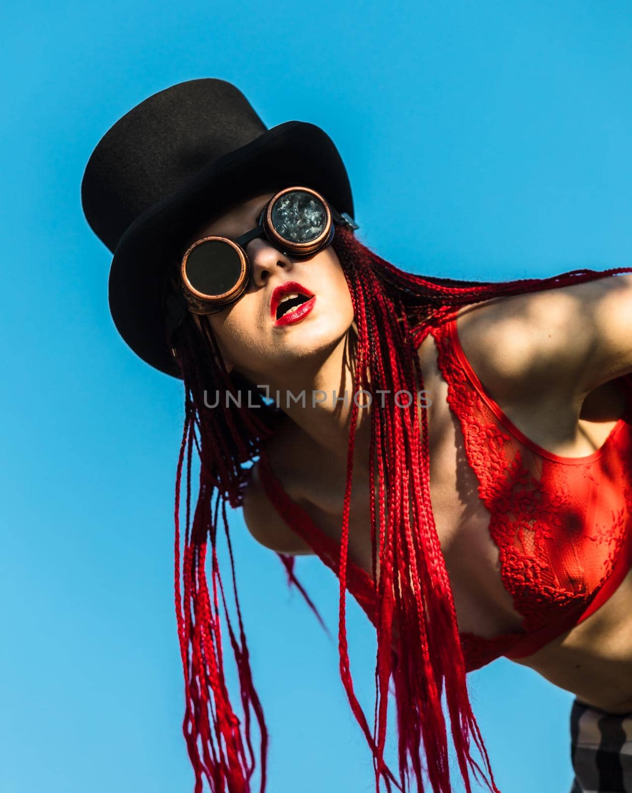 Glamorous girl with scarlet dreadlocks, a red swimsuit, black hat and welding glasses poses on the blue sky background. Free space for your text