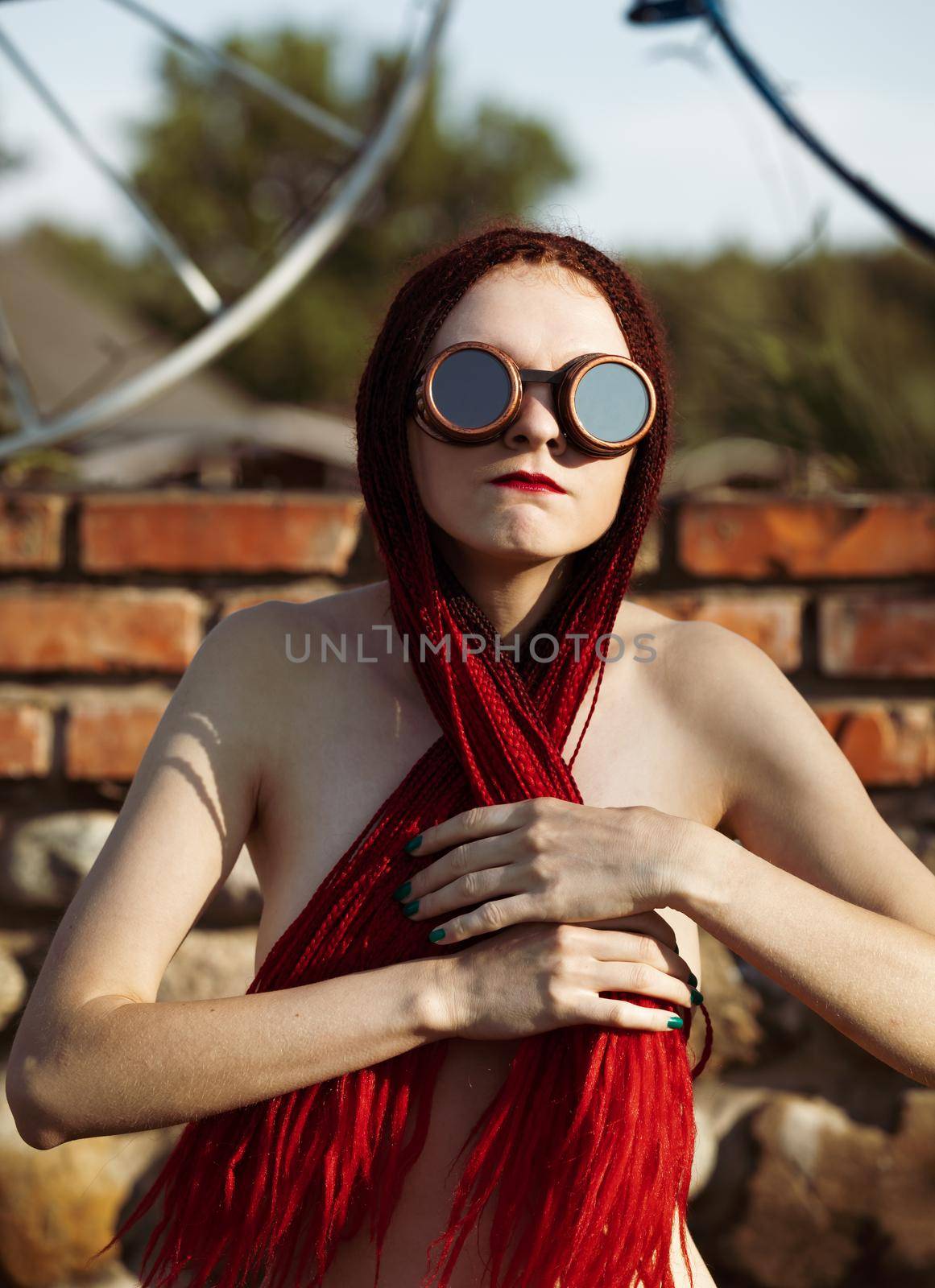 Semi-nude glamorous girl with scarlet dreadlocks and welding glasses closes the breast with scarlet dreadlocks