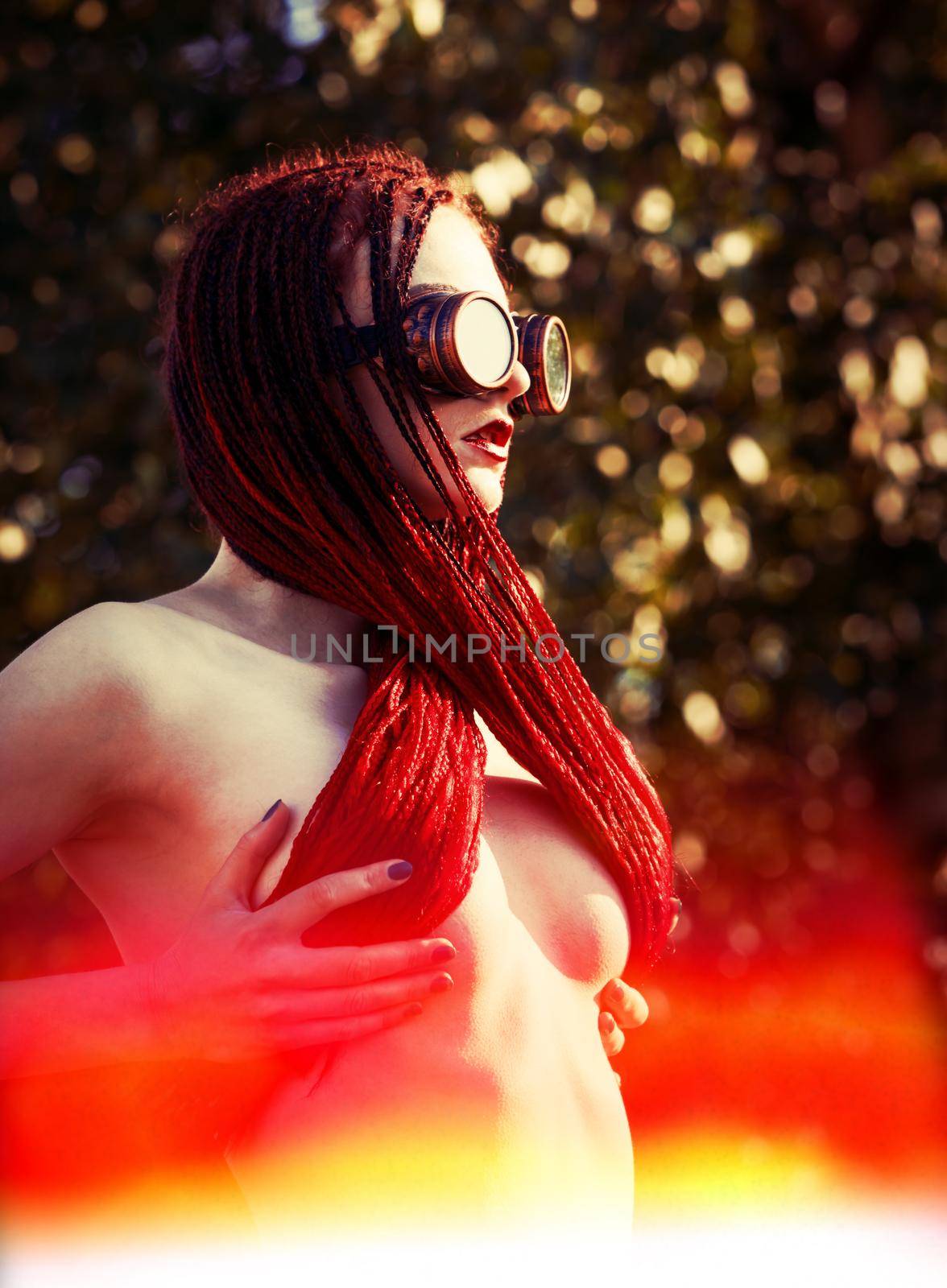 Semi-nude glamorous girl with scarlet dreadlocks and welding glasses closes the breast with scarlet dreadlocks